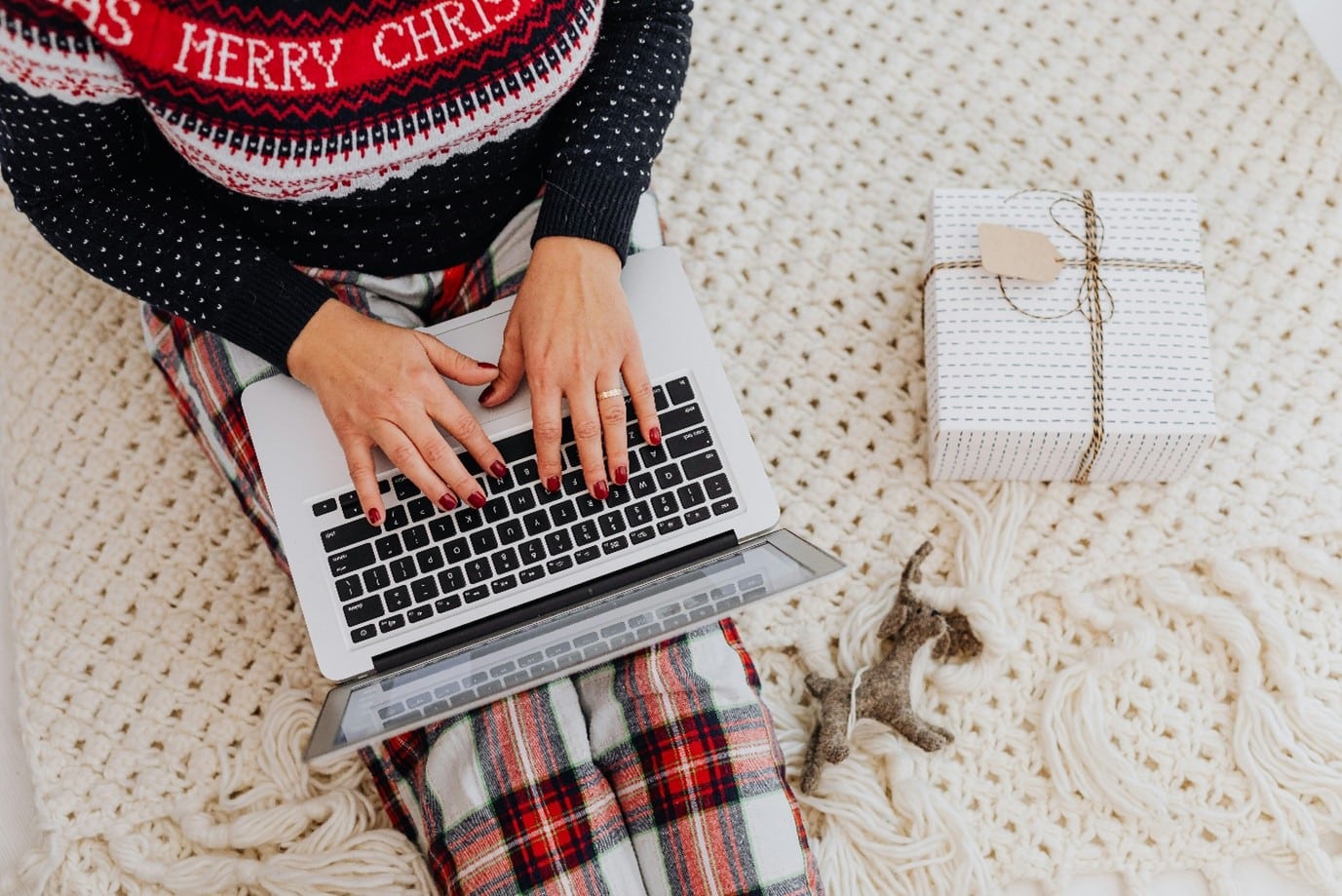 A person is wearing a festive Christmas outfit and typing on a laptop, sitting on a knitted blanked. A wrapped gift is next to a person.