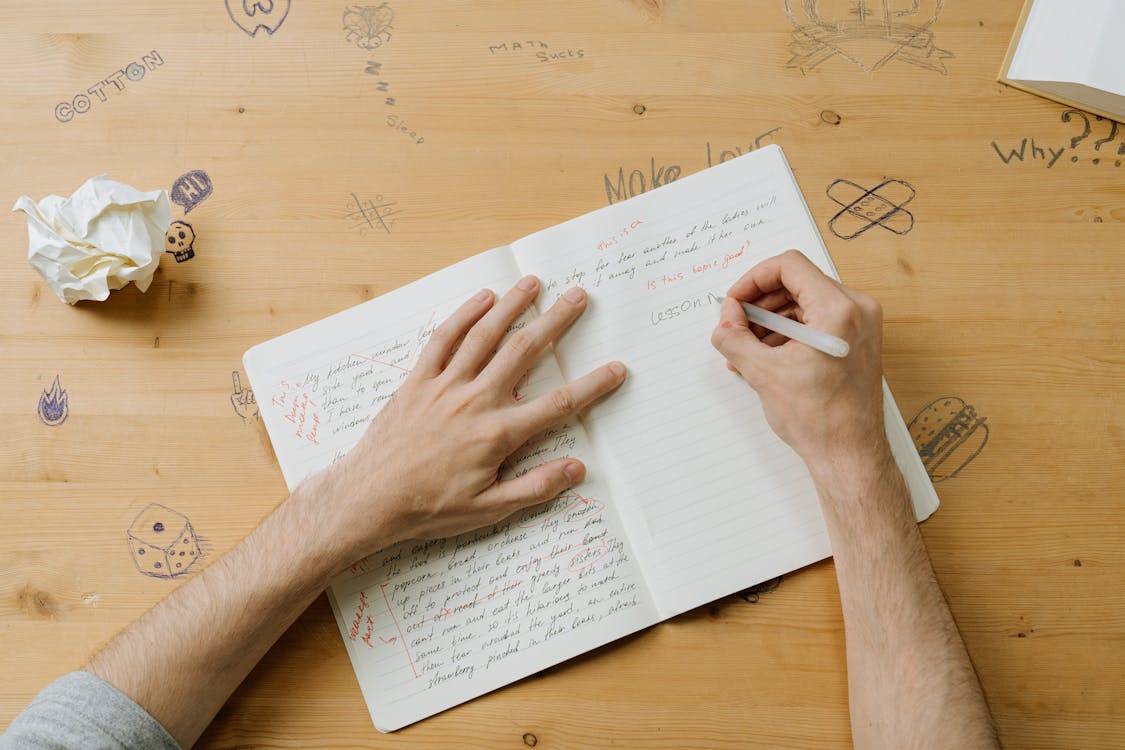 Free A person writing notes on a notebook with doodles on a wooden desk, reflecting creativity and education. Stock Photo